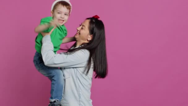 Mother lifting up her little boy and kissing him in studio, showing love and hugging cute kid against pink background. Sweet mom and son embracing each other and laughing. Camera A. - Footage, Video