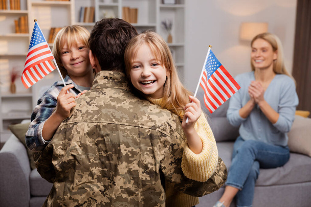 Heureuse famille américaine célébrant le retour d'un soldat. Père en tenue militaire, embrassant ses joyeux enfants, qui le saluent avec des drapeaux américains. Dans leur salon, mère excitée attend sur le canapé - Photo, image