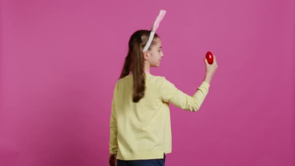 Enthusiastic small child spinning around in front of camera, presenting her red painted egg for easter sunday festivity. Cheery toddler with bunny ears twirling and doing a pirouette. Camera B. - Footage, Video