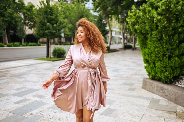 Die junge schöne Afroamerikanerin genießt einen Spaziergang im öffentlichen Park. Eine glückliche Frau plus Größe in seidenrosa Kleid lächelt durch die Straßen der Stadt - Foto, Bild
