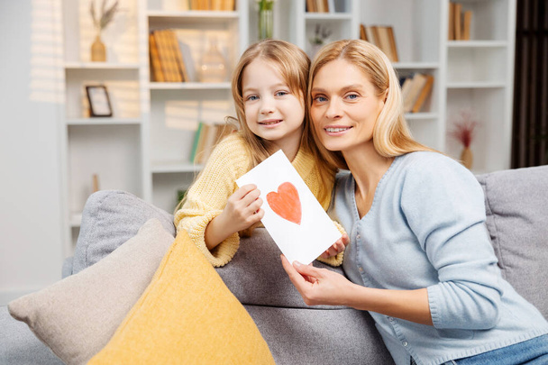 Happy family celebrating Mother's Day. A lovely mother and her young daughter with a handmade card looking at the camera, enjoying the moment on their sofa - Photo, Image