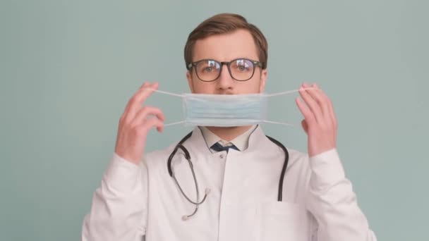 Gözlüklü ve steteskoplu bir doktor, yüzünde tıbbi bir maske tutuyor, gülümseme ve endişenin jestleri sırasında görme bakımının önemini gösteriyor.. - Video, Çekim