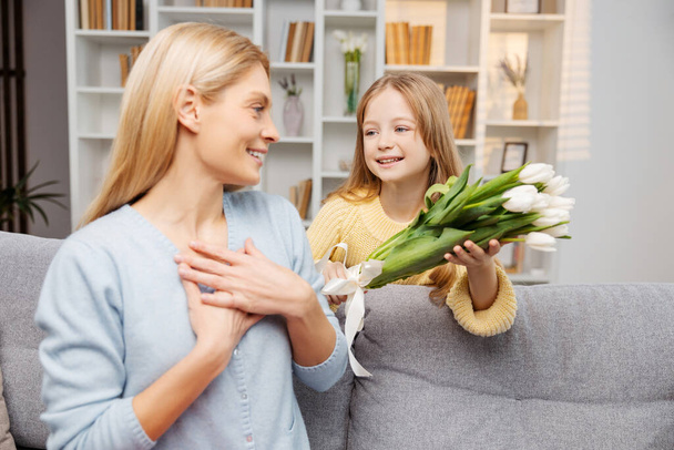 Happy family celebrating Mother's Day. Mother receiving tulips, surprised by her smiling daughter. Woman sitting on the living room sofa - Photo, Image