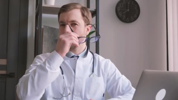 Een arts in een overhemd zit achter een bureau, terwijl hij zijn bril aanpast terwijl hij een laptop gebruikt voor gezichtsverzorging. Hij kan ook een stethoscoop in de buurt hebben.. - Video