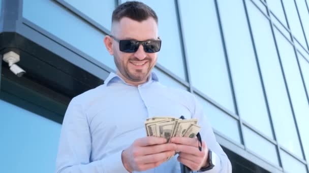 Un hombre con gafas de sol sonríe mientras sostiene un fajo de dinero en su mano cerca de un edificio de vidrio. Un hombre de negocios rico. Concepto de éxito - Imágenes, Vídeo
