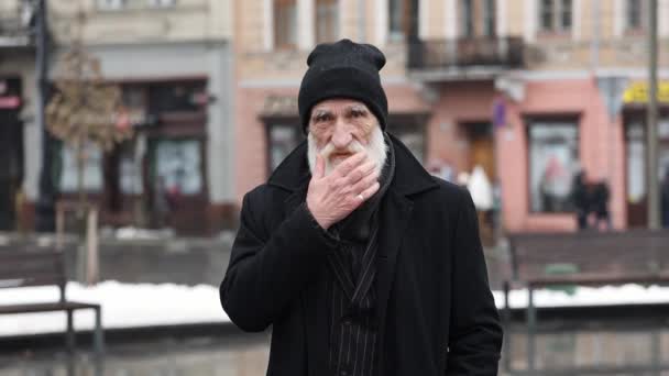 Sakalını okşayan ve kameraya bakan buruşuk sakallı yaşlı bir adamın portresi. Şehir binalarının arka planında, kışlık siyah ceketli pozitif yaşlı büyükbaba.. - Video, Çekim