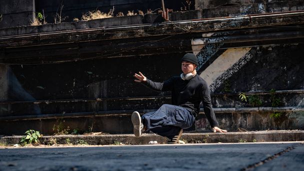 Um energético e profissional asiático hiphop breakdancer masculino em roupas da moda é breakdancing na rua abandonada, apreciando o ritmo. - Foto, Imagem