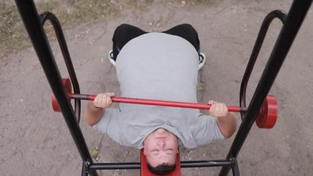Un hombre gordo con una camiseta gris levanta pesas mientras está acostado en una máquina de ejercicios al aire libre. Lucha con el exceso de peso. Levantando la barra - Imágenes, Vídeo