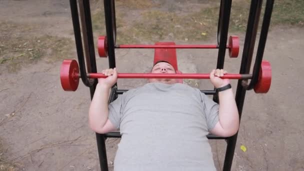 Un uomo grasso persistente solleva pesi sdraiati su macchine per l'esercizio all'aperto. Lotta con il peso in eccesso. Addestramento uomo sullo sfondo di asfalto - Filmati, video
