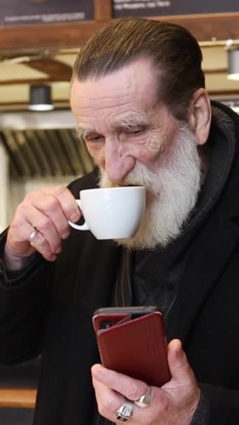 Vanha ryppyinen mafioso mustissa vaatteissa katsoo puhelinta ja juo kupin kahvia kahvilassa. Rikas isoisä. Rikkaat vanhukset - Materiaali, video