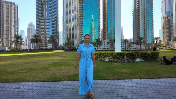 Mujer feliz en traje azul de verano caminando en el parque de la ciudad. Acción. Pradera verde y rascacielos en el fondo - Foto, imagen