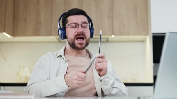 Portret ekspresyjnego mężczyzny w bezprzewodowych słuchawkach udającego, że gra na perkusji z ołówkami w rękach w jasnym mieszkaniu. energiczny facet słuchając online streaming muzyka i ciesząc się bas wewnątrz. - Materiał filmowy, wideo