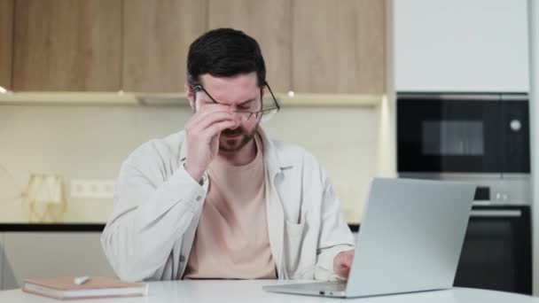 Hombre de estilo casual sobrecargado quitándose los anteojos y frotándose los ojos mientras trabaja por computadora portátil en el espacio de trabajo doméstico. Tipo estresado que experimenta problemas de visión después de mirar fijamente a la pantalla durante mucho tiempo. - Metraje, vídeo