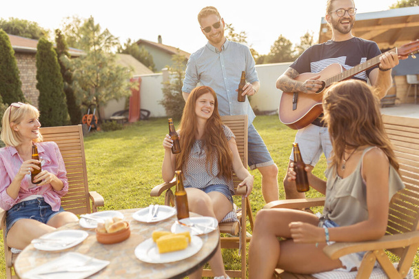 Group of friends having fun playing the guitar and singing at backyard barbecue party, drinking beer and relaxing outdoors on a sunny summer day - Fotografie, Obrázek