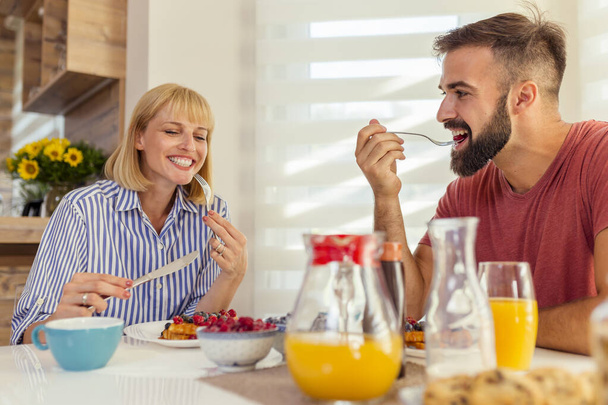 Beautiful young couple in love sitting at dinning room table, having waffles, berries, orange juice and coffee for breakfast, spending weekend morning together at home - Φωτογραφία, εικόνα