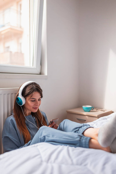 Woman sitting on the floor by the bed in bedroom, relaxing at home in the morning, wearing headset and learning foreign language using online smart phone app - Foto, Bild