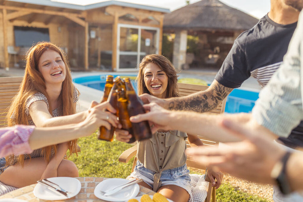 Group of cheerful young friends having fun at poolside backyard barbecue party, making a toast with bottles of beer and enjoying sunny summer days outdoor - Photo, Image