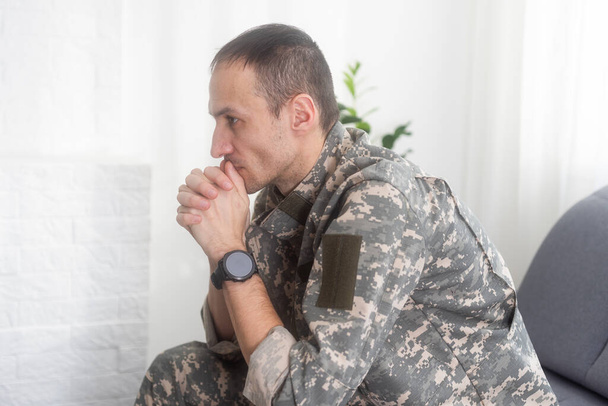 Портрет спокойного серьезного кавказского военнослужащего в камуфляжной форме и кепке, сидящего на диване, с депрессивным выражением лица. Высокое качество фото - Фото, изображение