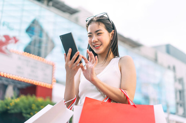 Lidé městský životní styl s nákupem spotřební zboží. Mladý dospělý asijské žena pomocí smartphone psaní zprávy sociální média. Šťastný úsměv tvář stojící venku. - Fotografie, Obrázek