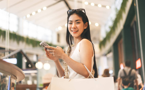 La gente de la ciudad estilos de vida con la compra de consumismo de compras. Joven adulto asiático mujer usando smartphone. Cara de sonrisa feliz sentado en los grandes almacenes interiores. - Foto, Imagen