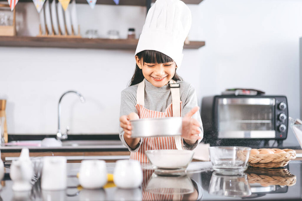 Маленькая азиатская девочка в шляпе шеф-повара и фартуке, готовящая на кухне. Подготовка к домашнему торту. Досуг образ жизни с дома. Рост ребенка с интересной концепцией. - Фото, изображение