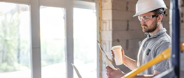 Een elektricien bestudeert een bouwtekening met een koffie in zijn hand. Kopieerruimte. - Foto, afbeelding