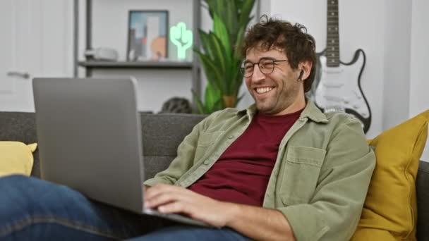 Komea mies nauraa käyttäen kannettavaa tietokonetta sohvalla modernissa olohuoneessa. - Materiaali, video