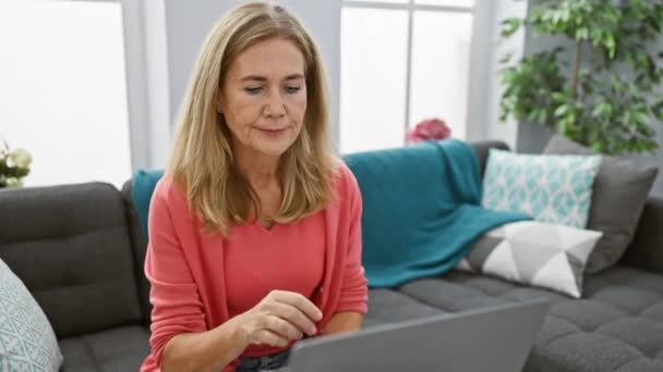 Reife blonde Frau betet zu Hause vor ihrem Laptop in einem gemütlichen, modernen Wohnzimmer. - Filmmaterial, Video