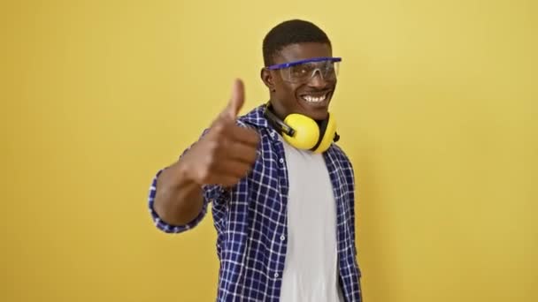 Cheery african american man, in safety glasses, confidently stands, flashes an excellent approval sign, joy evident in his smile. isolated against a vibrant yellow background, he embodies success. - Footage, Video
