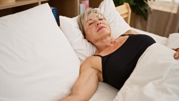 Mujer serena de mediana edad con el pelo gris relajante en el dormitorio acostado en ropa de cama blanca en el interior - Metraje, vídeo
