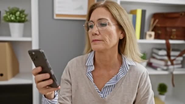 Starší podnikatelka v brýlích pomocí chytrého telefonu v moderním kancelářském interiéru, vyjadřující obavy a spokojenost. - Záběry, video