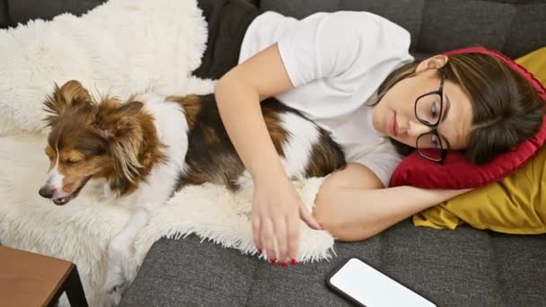 Fiatal nő szemüvegben feküdt mellett alvó kutya kanapén otthon sms-ezés közben - Felvétel, videó