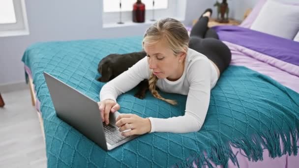 Una donna bionda si trova su un letto con un computer portatile, il suo cane labrador riposa accanto a lei in un ambiente accogliente camera da letto. - Filmati, video