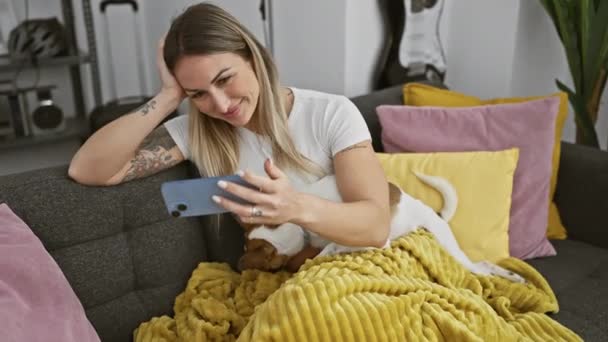 Een glimlachende vrouw ontspant met haar hond op de bank terwijl ze een smartphone gebruikt in een moderne woonkamer. - Video