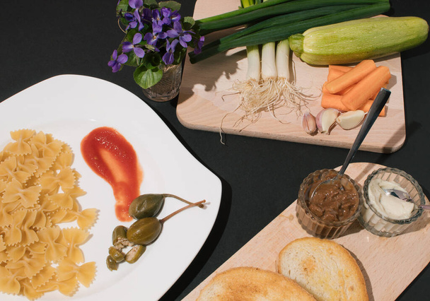 Kuchyňský stůl s talířem, dřevěnými deskami, zeleninou, krajíčkem chleba, těstovinovou mašlí a fialkami. Koncept zdravé stravy. - Fotografie, Obrázek
