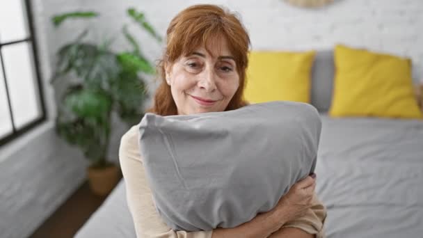 Hymyilevä keski-ikäinen nainen istuu sängyllään, halaa tyynyä viileällä, onnellisella hymyllä, näyttää hampaita makuuhuoneessaan, säteilee luottamusta, iloa ja luonnon kauneutta. - Materiaali, video