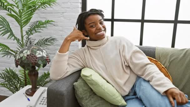 Joven y alegre mujer negra con rastas haciendo un gesto amistoso de llamada telefónica mientras está sentada en un sofá en casa - Metraje, vídeo