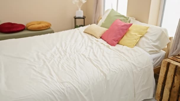Молодая женщина в розовой пижаме организовывает стирку в плетеной корзине на кровати с красочными подушками в уютной спальне. - Кадры, видео
