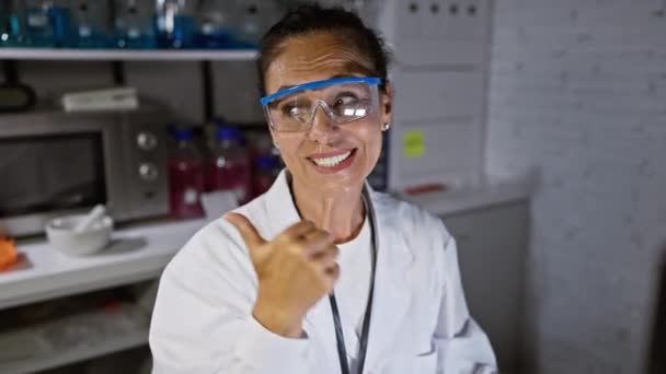 Vidám középkorú spanyol tudós nő, biztonsági szemüveggel, boldogan mutat a számítógép oldalára, hüvelykujjával felfelé mutat a laborban, nyitott szájjal mosolyog, örömöt sugároz.! - Felvétel, videó