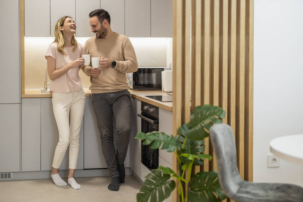 Junger Mann und Frau stehen in der Küche und reden. Fröhliche Frau und Mann mit Tassen Kaffee. - Foto, Bild