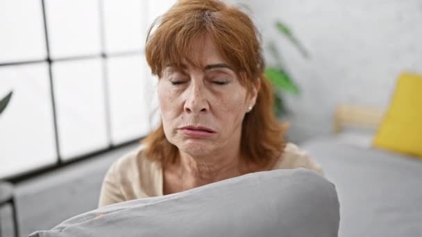 Sceptyczna kobieta w średnim wieku, negatywna osoba, siedzi nerwowo na łóżku, przytulając poduszkę, marszcząc brwi zdenerwowanymi emocjami nad problemem, w sypialni - Materiał filmowy, wideo