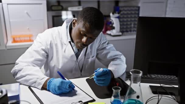 Afrikaanse mannelijke wetenschapper maakt notities in een laboratorium omgeving, met testapparatuur en een computer op de achtergrond. - Video
