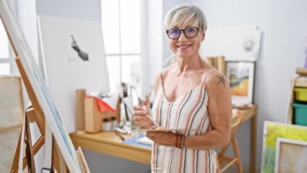 Χαμογελώντας ώριμη γυναίκα ζωγραφική σε καμβά σε ένα φωτεινό στούντιο τέχνης - Πλάνα, βίντεο