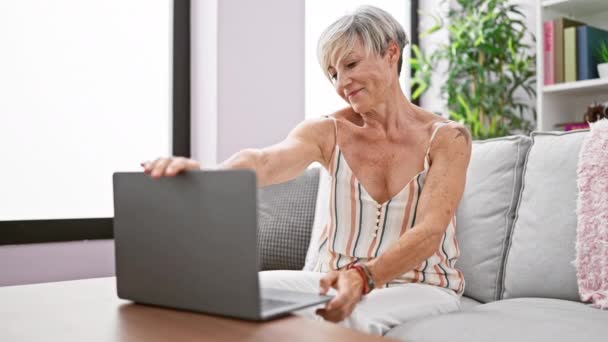 Елегантна старша жінка використовує ноутбук на дивані вдома, випромінюючи впевненість і комфорт у своєму житловому просторі. - Кадри, відео