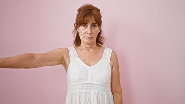Ώριμη, θυμωμένη γυναίκα με ροζ φόρεμα κάνει χειρονομίες προς τα κάτω, μια εικόνα απόρριψης και δυστυχίας, απομονωμένη σε ροζ φόντο - Πλάνα, βίντεο