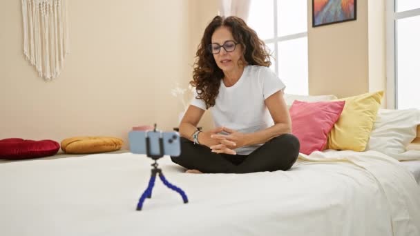 Mulher hispânica madura com cabelo encaracolado sentado de pernas cruzadas em sua cama em um ambiente confortável em casa, gravando com um smartphone em um tripé. - Filmagem, Vídeo