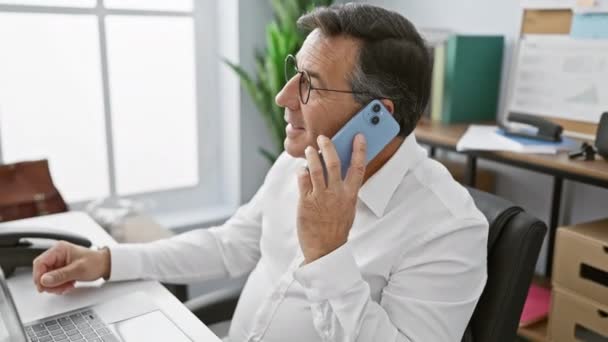 Homme d'affaires mature en lunettes utilisant un smartphone dans un cadre de bureau moderne - Séquence, vidéo