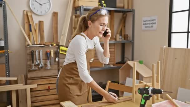 volwassen blonde vrouw in schort praten op telefoon en markeren van hout in een goed uitgeruste werkplaats - Video