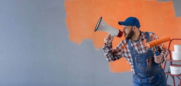 Красивый бородатый художник с роллером, громко кричащий мегафоном, стоящим возле стены в комнате, делая ремонт дома. Концепция маркетинга или продаж. баннер. Копирование пространства - Фото, изображение