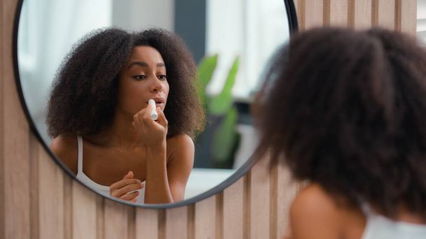 浴室の浴室の浴室の鏡の反射 アフリカ系アメリカ人の美女のメイクアップ 自然な滑らかな保湿口紅の女性の少女は化粧品の唇の光沢の笑顔を保湿する唇のバームのスキン ケアを適用します - 写真・画像